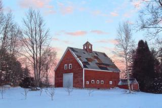 A weathered red barn amidst winter trees and a snow covered field at dusk.